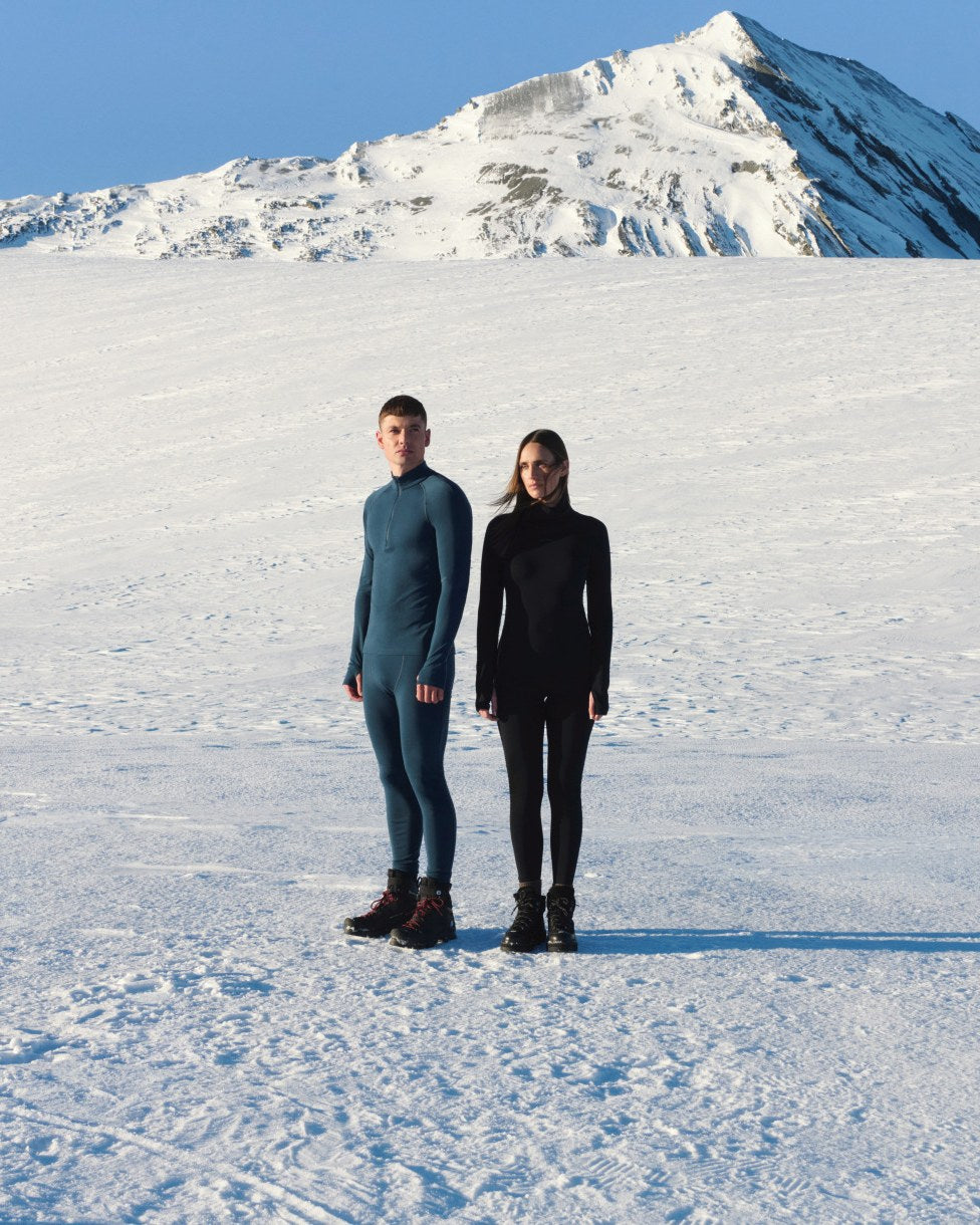 Two people in icebreaker merino base layers standing on snow below a mountain peak