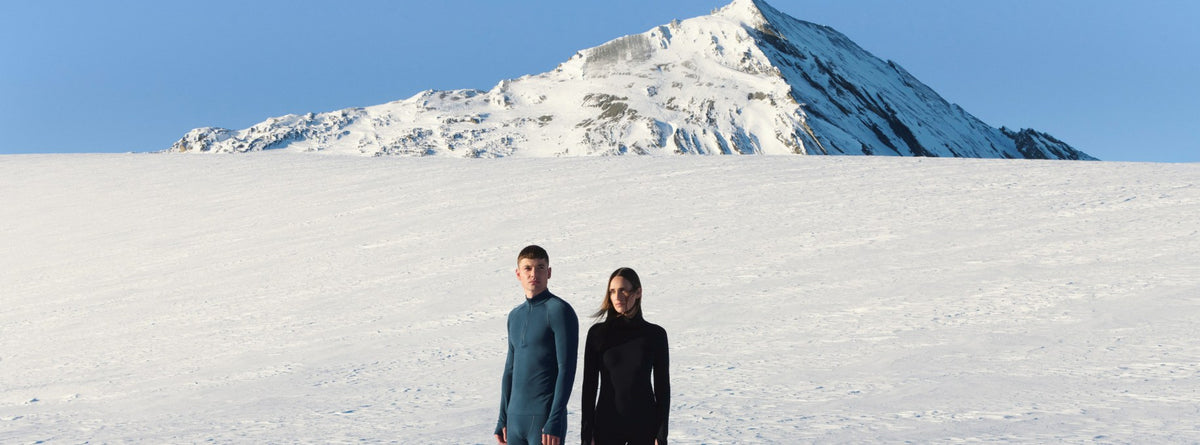 Two people in icebreaker merino base layers standing on snow below a mountain peak
